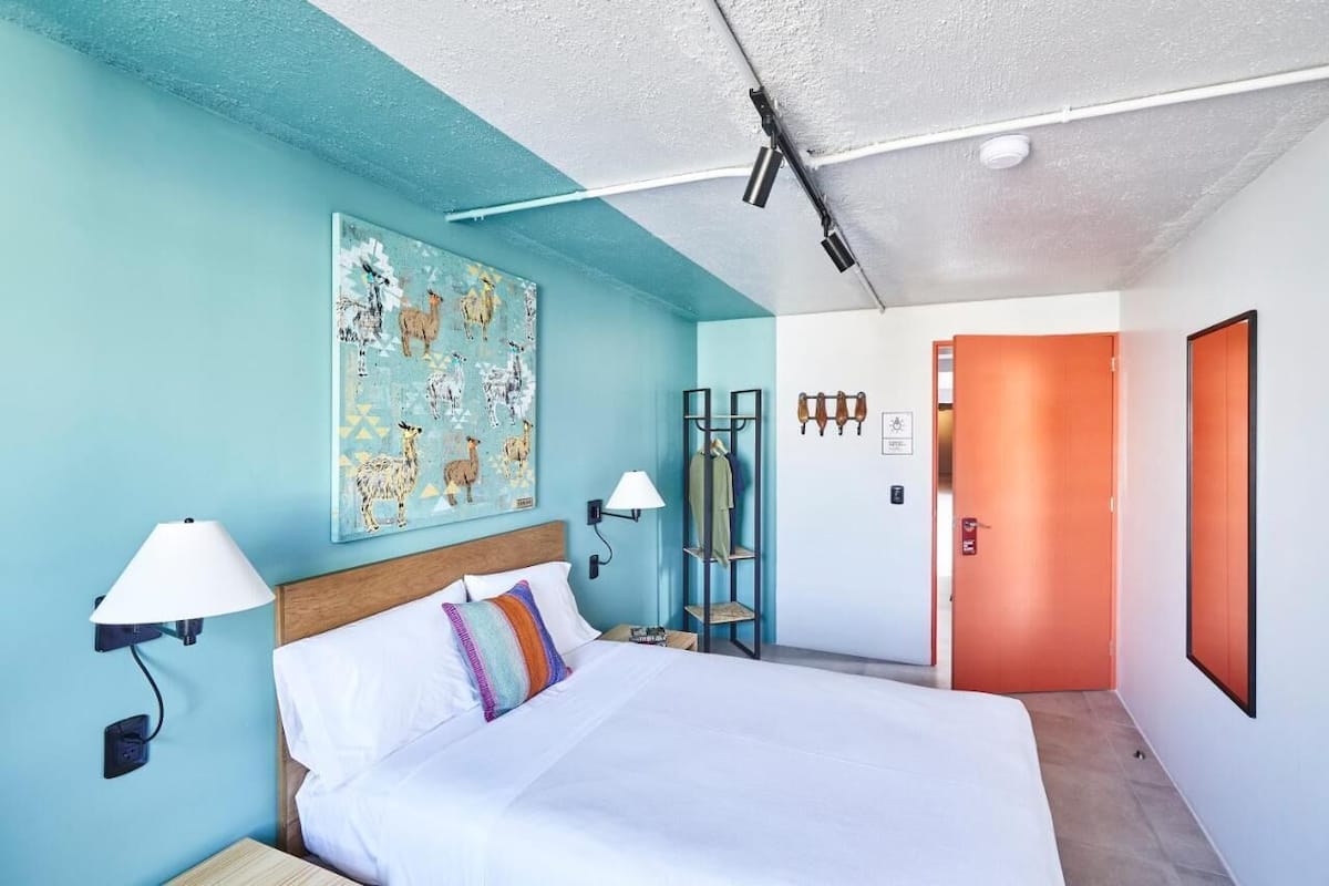 A hotel room with a bed, a nightstand, a wall-mounted lamp, a painting, a wardrobe, and an orange door.
