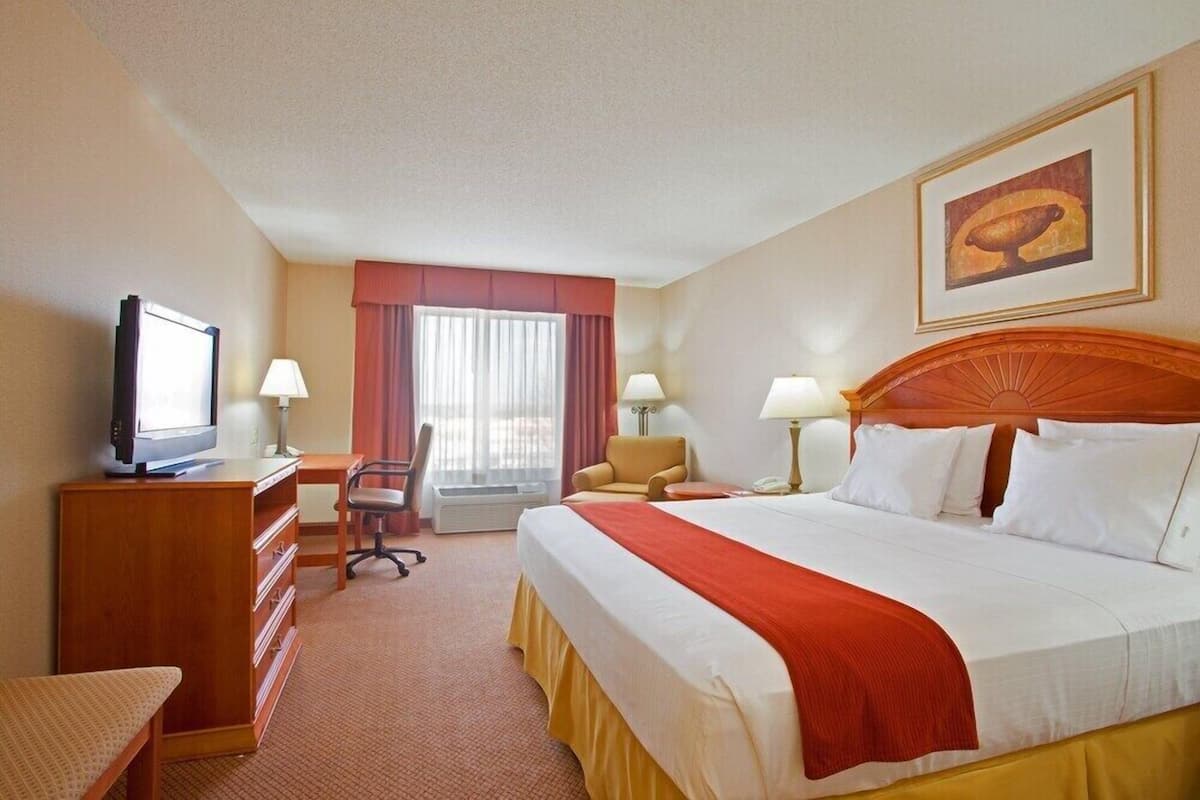 A hotel room with a bed, a desk with a television, a chair, a desk lamp, and a window with curtains.