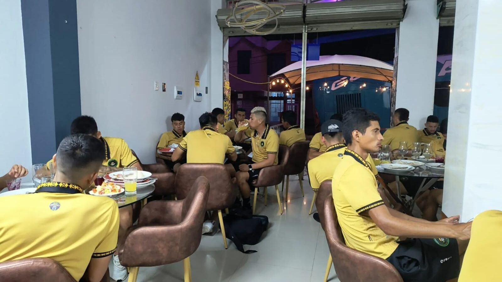 Un área de comedor con gente sentada en mesas, usando uniformes amarillos, en una habitación con paredes blancas y una pared de acento morada.
