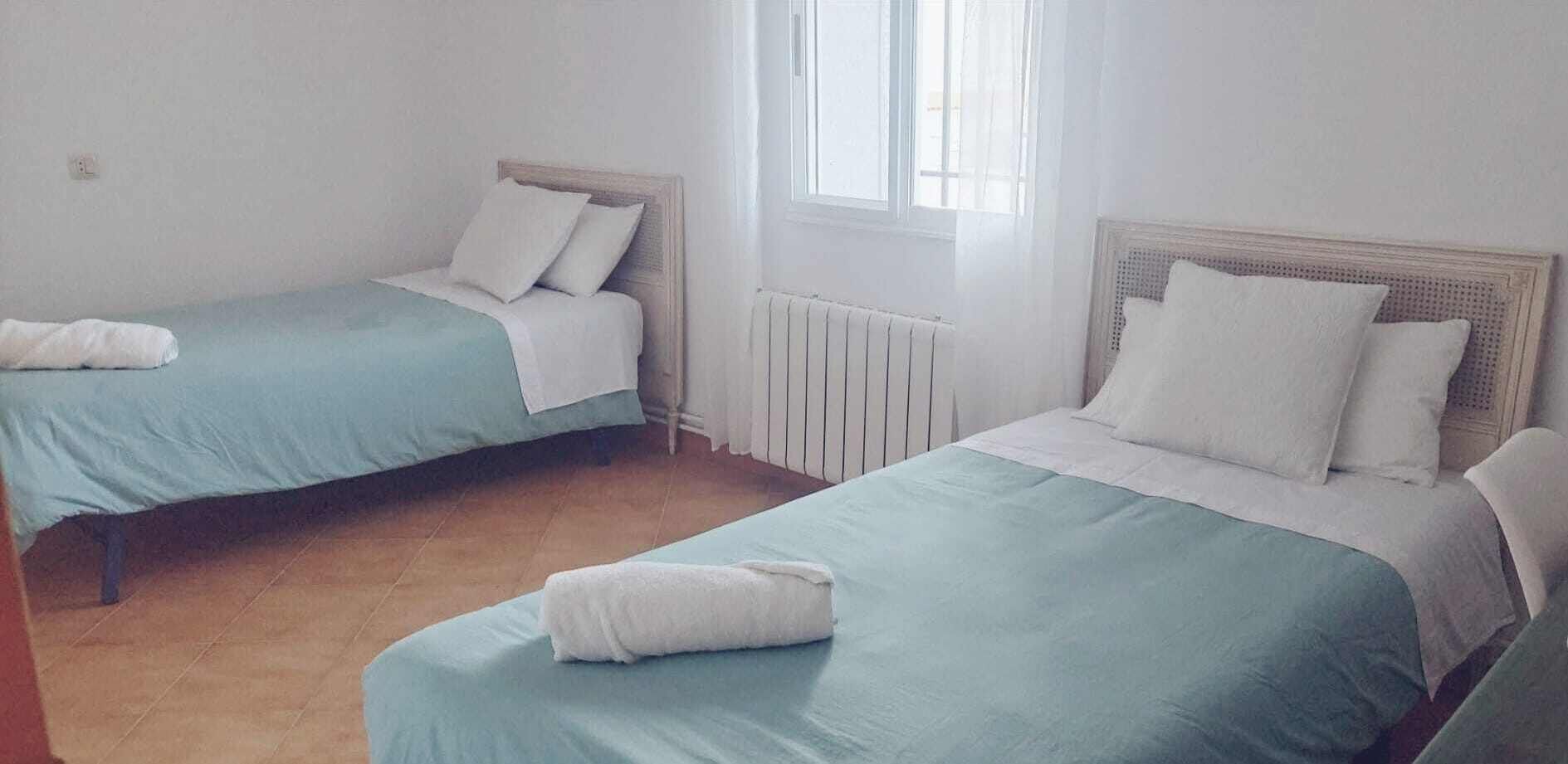 Two single beds with white pillows and blue bedding, a radiator, and a window with light coming through.