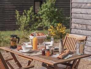 A wooden table set outdoors with a rustic setting, including a basket of bread, a bottle of juice, and a vase of yellow flowers.