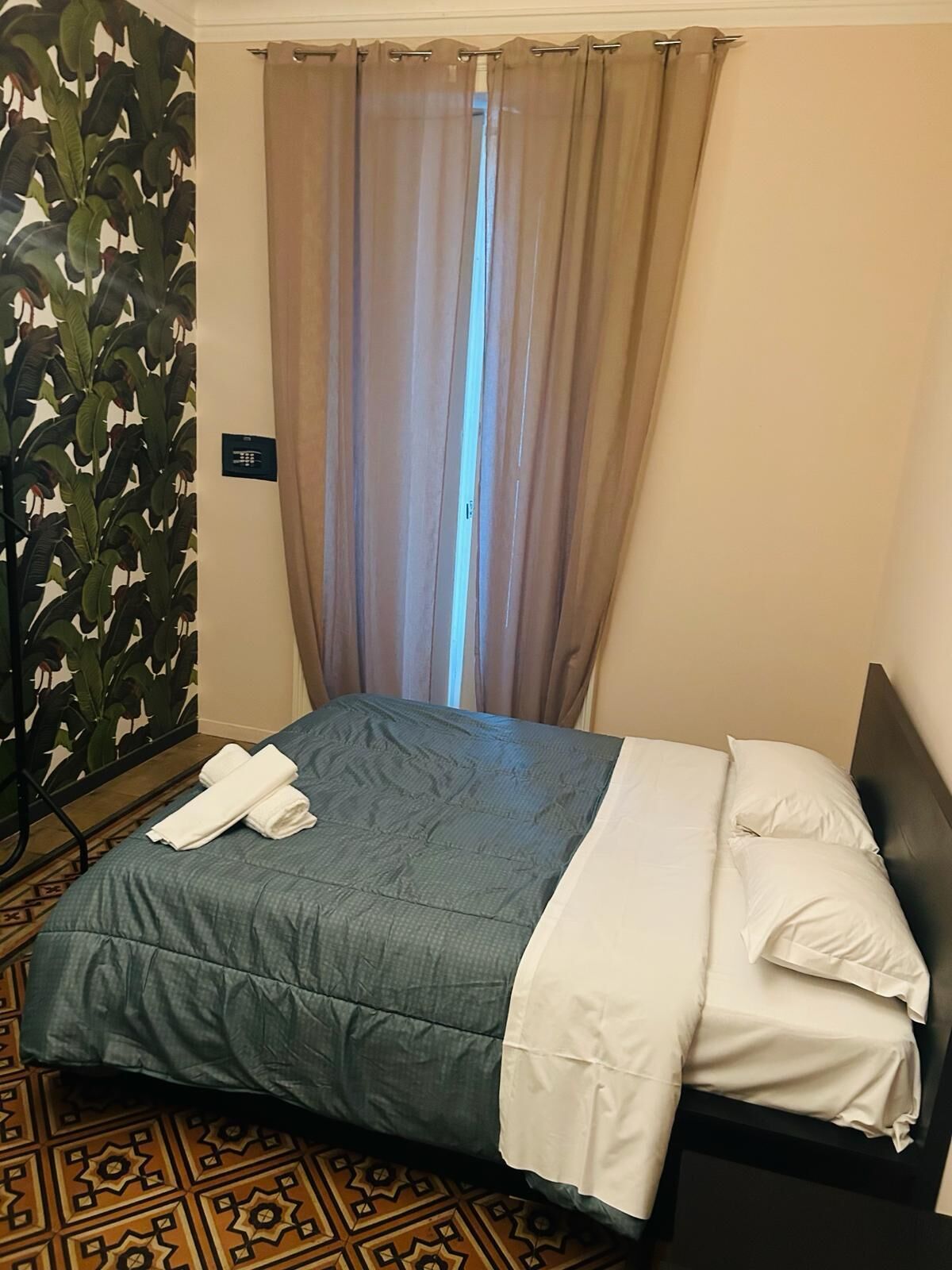 Una cama bien hecha con un edredón azul y blanco, un suelo estampado y una cortina cubriendo una ventana.