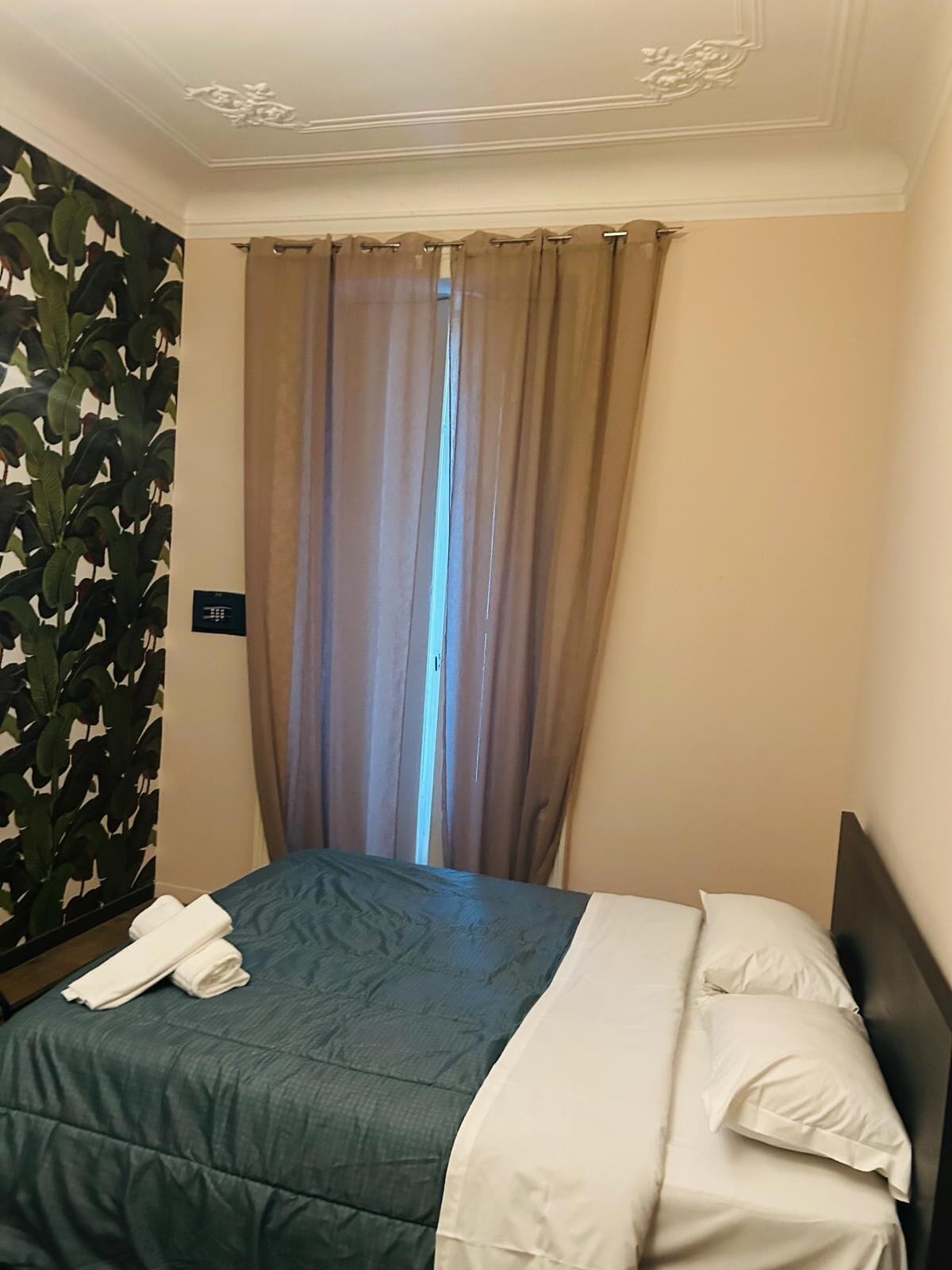 Een hotelkamer met een bed, beige gordijnen en een muur met een bladpatroon.