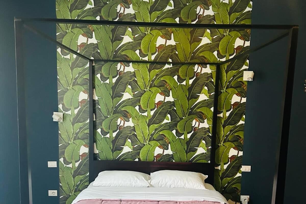 A four-poster bed with a pink bedspread and white pillows, set against a wall with a green leafy wallpaper.