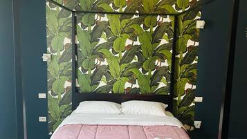 Una cama con dosel con un edredón rosa y almohadas blancas, frente a una pared con papel tapiz de hojas verdes.