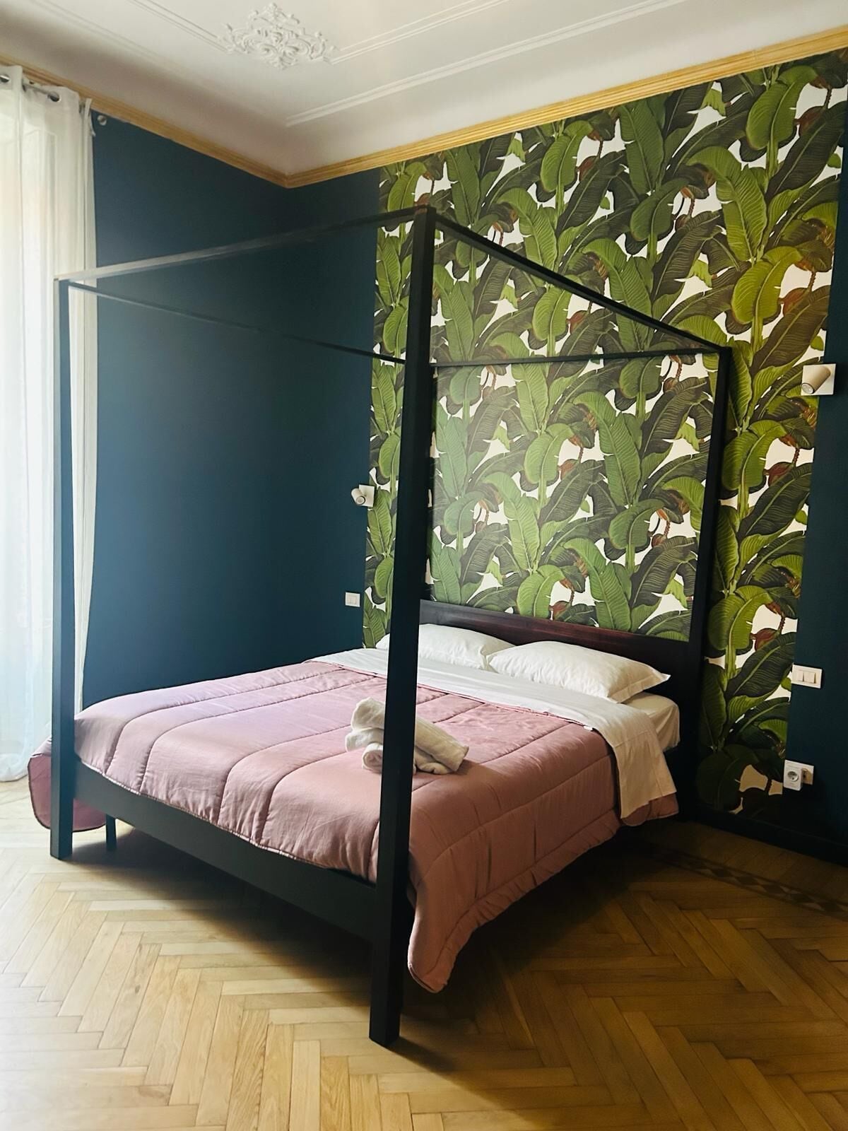 Ein Himmelbett mit rosa Bettdecke und weißen Kissen, vor einer Wand mit großem tropischen Blattmuster.