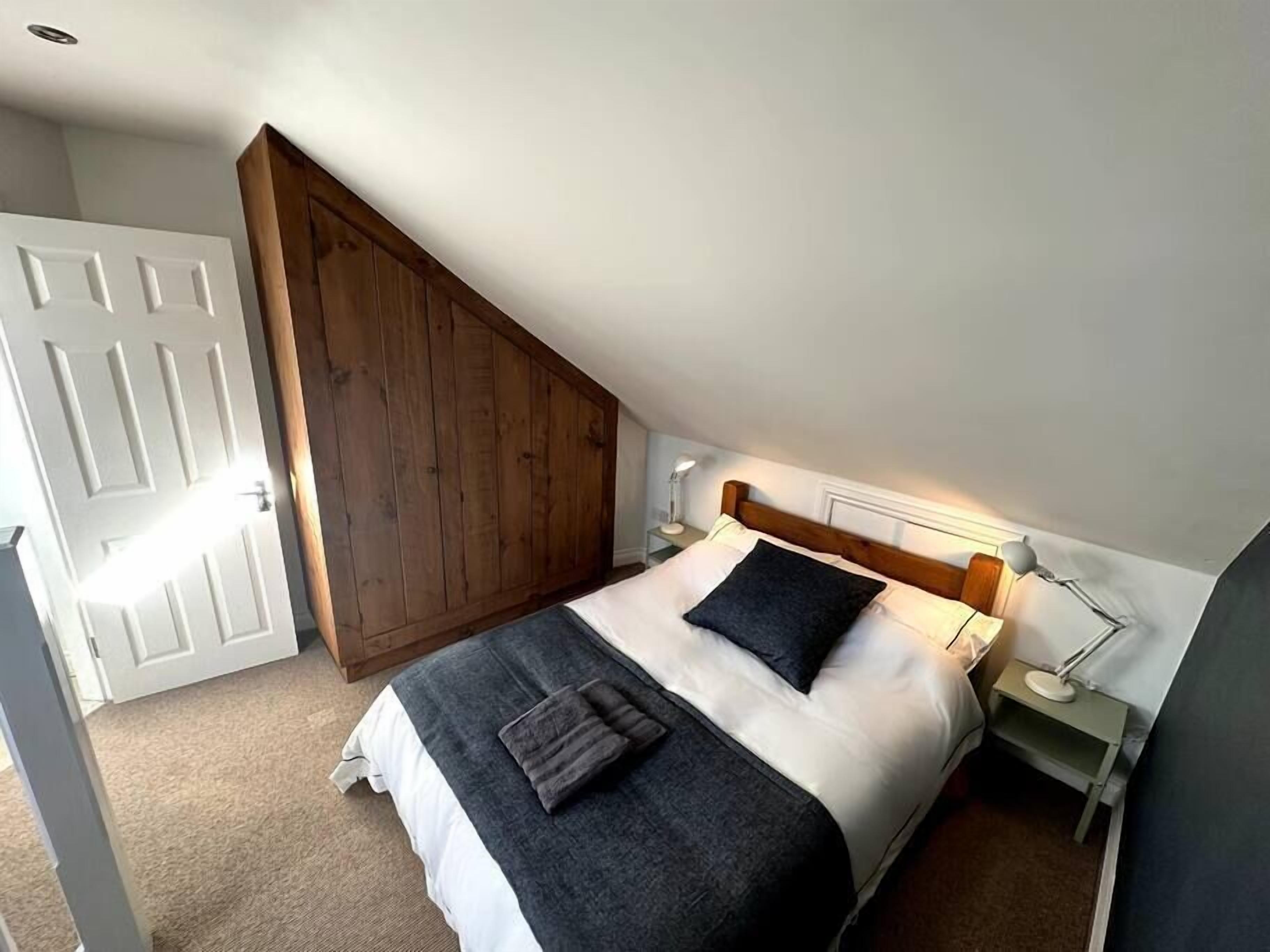 A bedroom with a sloped ceiling, a wooden wardrobe, a bed with white and dark bedding, and bedside lamps.