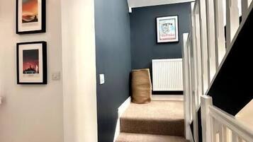 Een trap met beigekleurig tapijt en witte leuningen die uitkomt op een plateau met een radiator en afbeeldingen in lijsten aan de muur.
