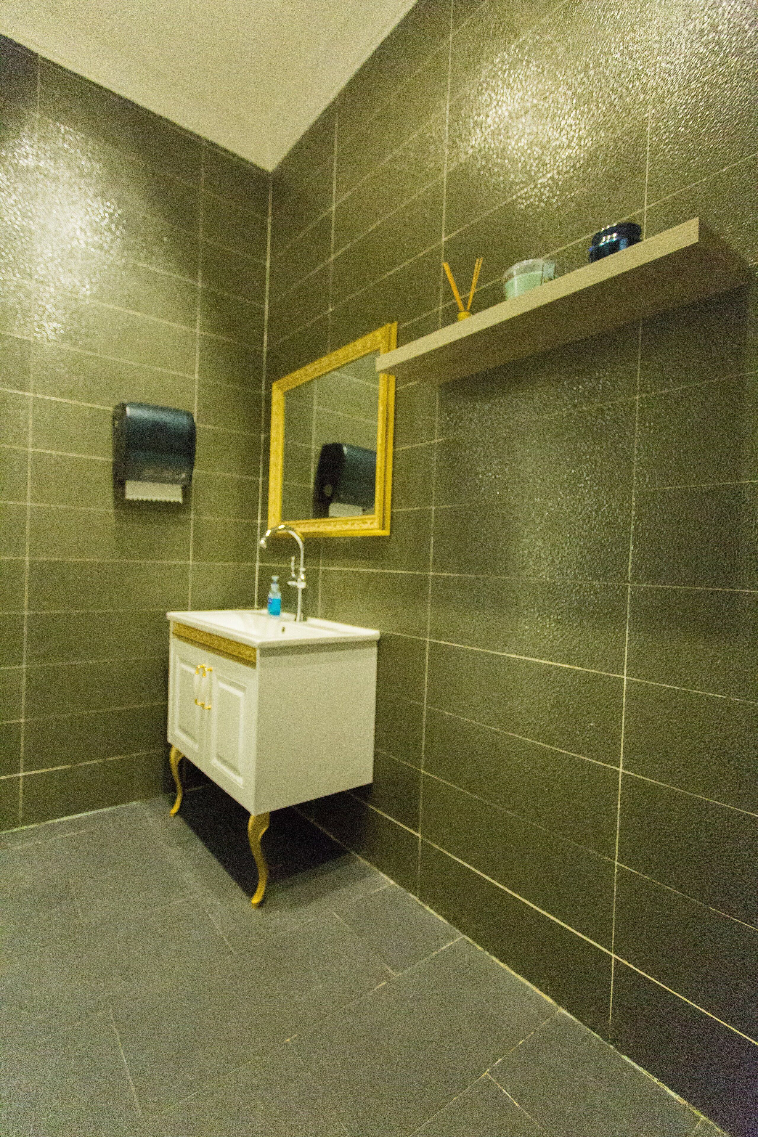 Ein Badezimmer mit dunkelgrünen Fliesen, einem weißen Waschbecken mit goldfarbenen Beinen, einem Spiegel mit goldener Rahmung und einem Ablagefach für Toilettenartikel.
