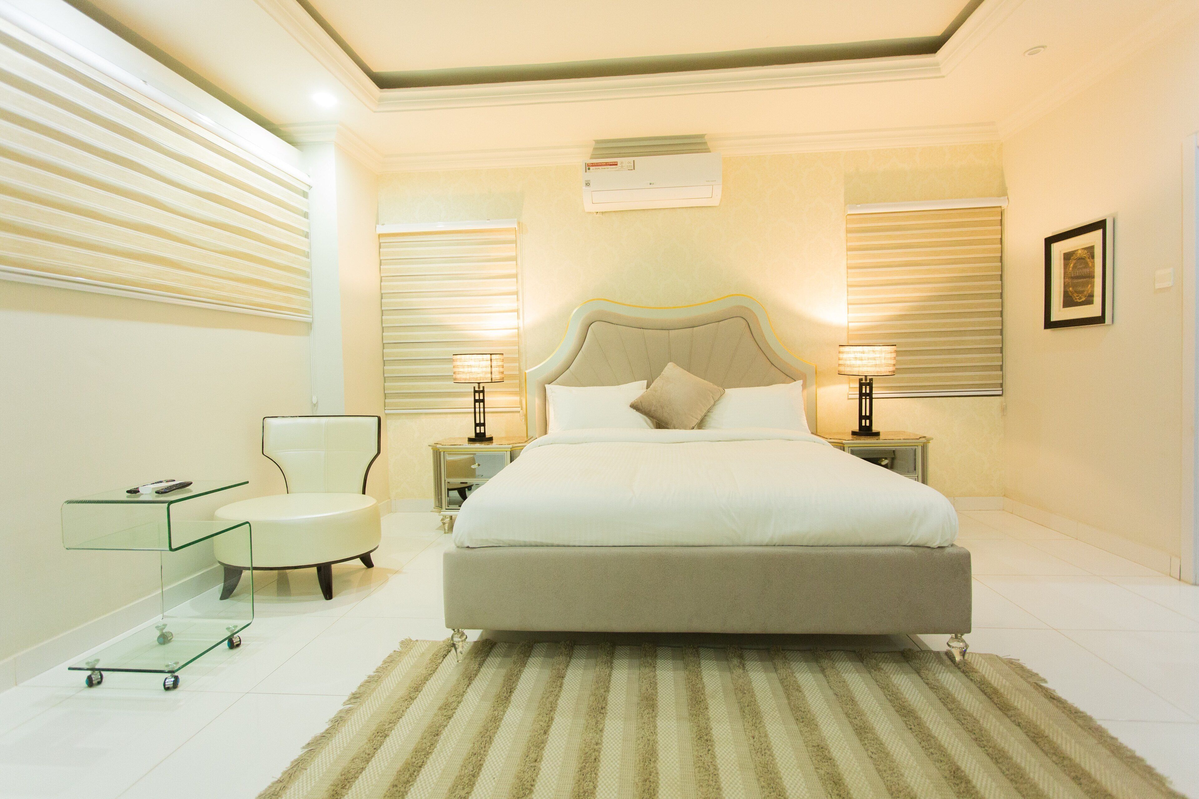 A modern bedroom with a large bed, bedside lamps, a glass cart, and a chair.