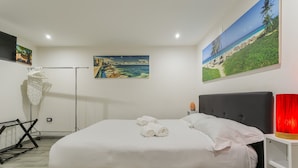 Un lit bien fait, avec du linge de lit blanc et deux serviettes enroulées, une tête de lit noire, une lampe rouge et deux tableaux représentant des paysages côtiers.