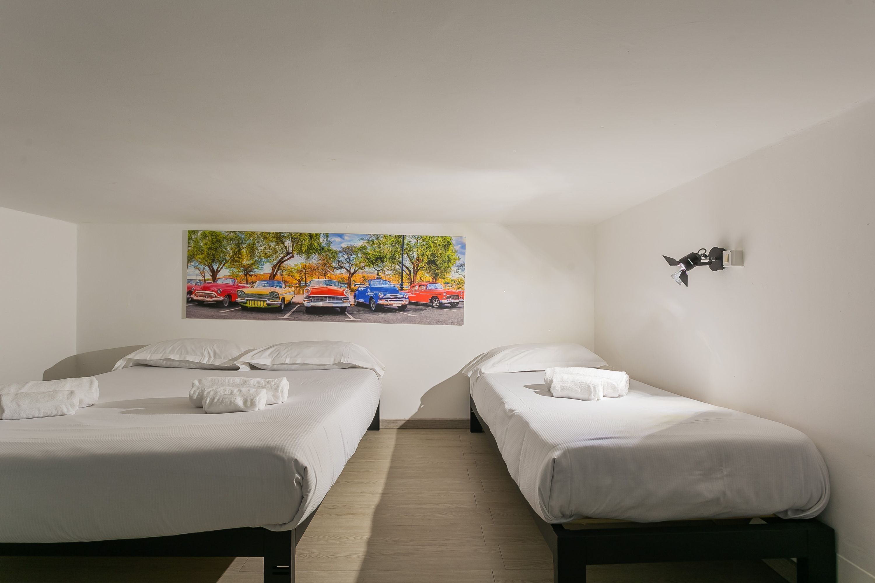 A hotel room with two single beds, a painting of a parking lot, and a wall-mounted light fixture.