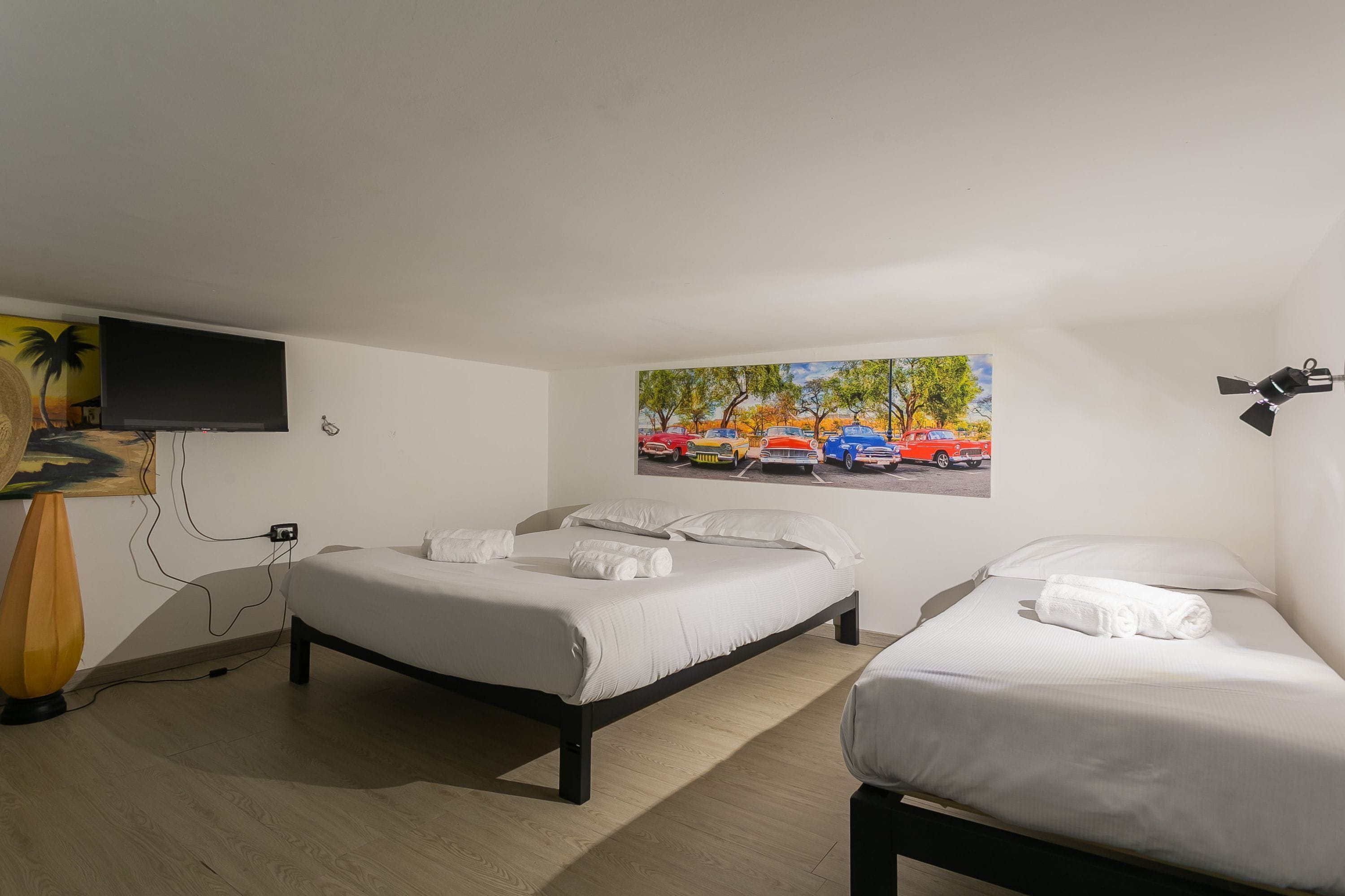 A room with two beds, a flat-screen TV mounted on the wall, a painting, and a ceiling fan.