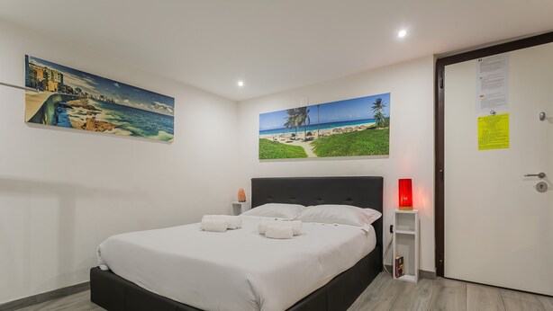 Un lit bien fait, avec du linge de lit blanc et des oreillers, une table de chevet avec une lampe, et deux tableaux accrochés au mur représentant des paysages côtiers.