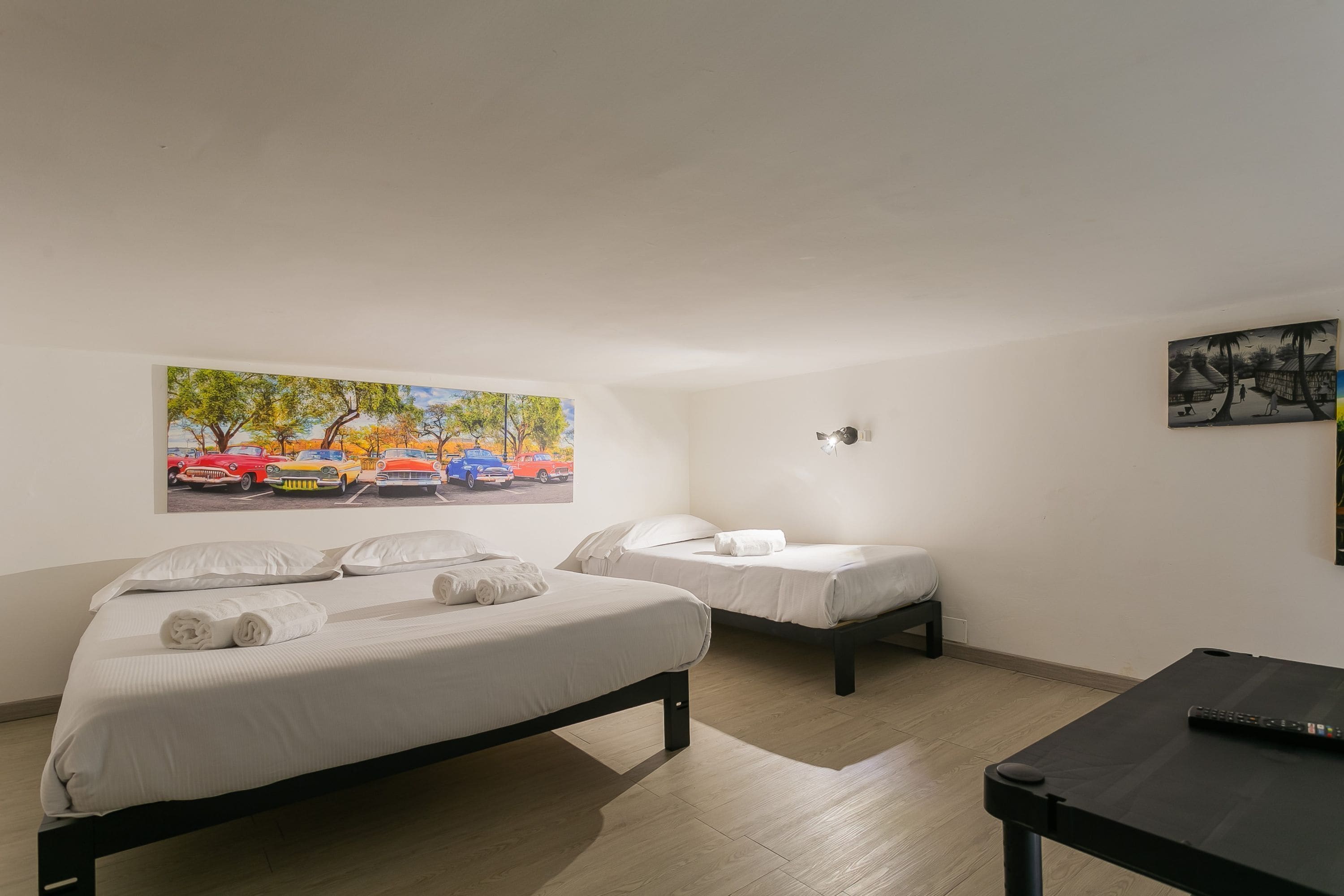 A hotel room with two beds, a desk, a painting of cars, and a wall-mounted light fixture.