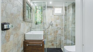 Une salle de bain avec un mur en pierre, un lavabo blanc, un meuble en bois, une cabine de douche en verre et des toilettes.