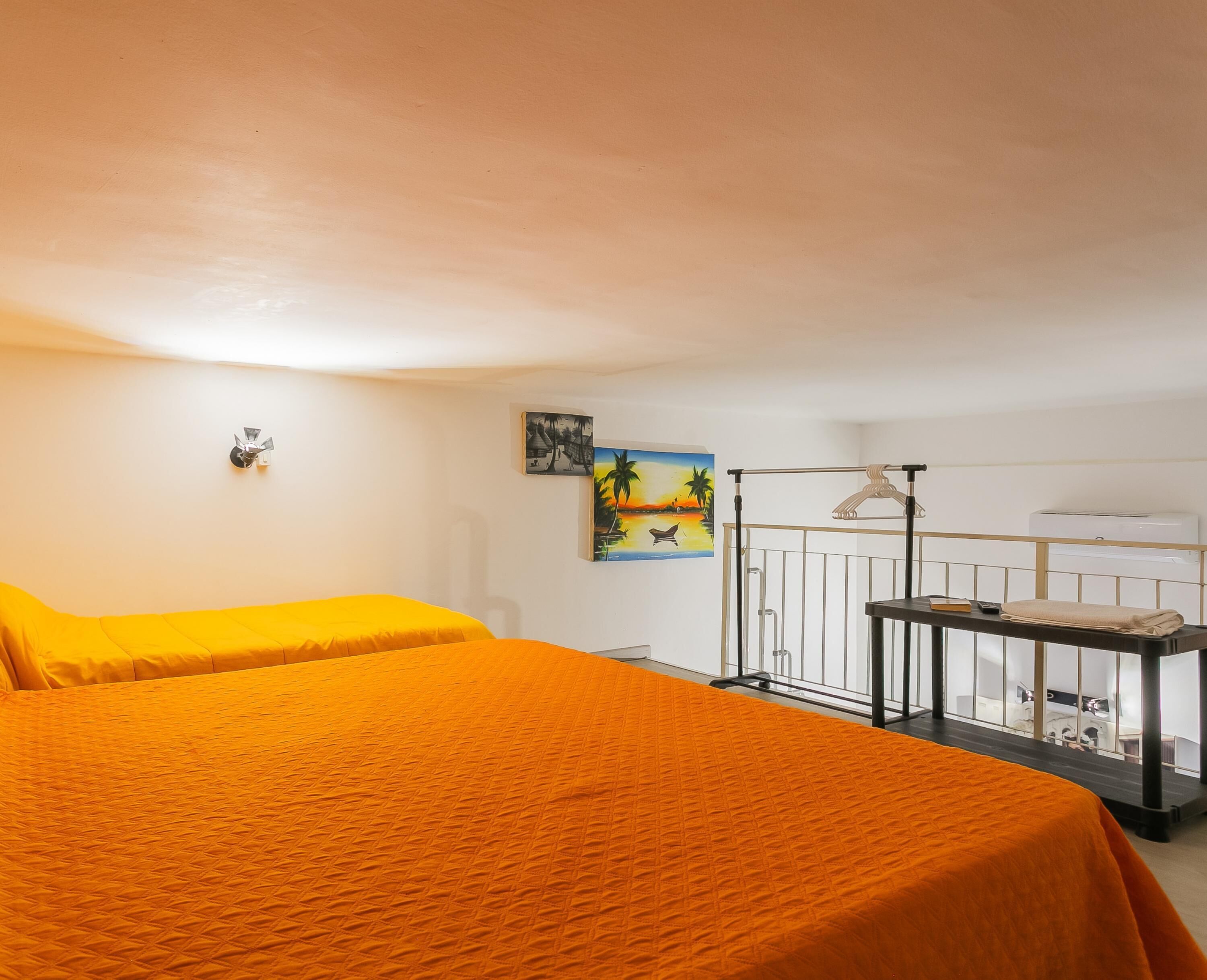 Ein Zimmer mit zwei Betten, einem Gemälde von einer Strandlandschaft, einem kleinen Tisch mit einem Buch und einem Handtuch sowie einem Balkon mit Geländer.