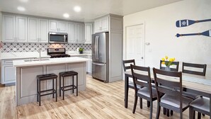 Une cuisine moderne avec des meubles blancs, un îlot central doté de tabourets, un réfrigérateur en acier inoxydable et un coin repas avec des chaises en bois et un vase de fleurs.