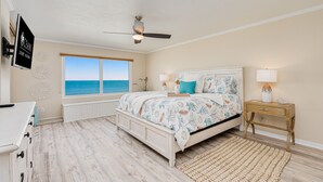 A bedroom with a large window, a ceiling fan, a bed with floral bedding, a bedside table with a lamp, and a white dresser. - NEW!Pool!Hot Tub!Oceanfront! Peninsula #303 (Daytona Beach)