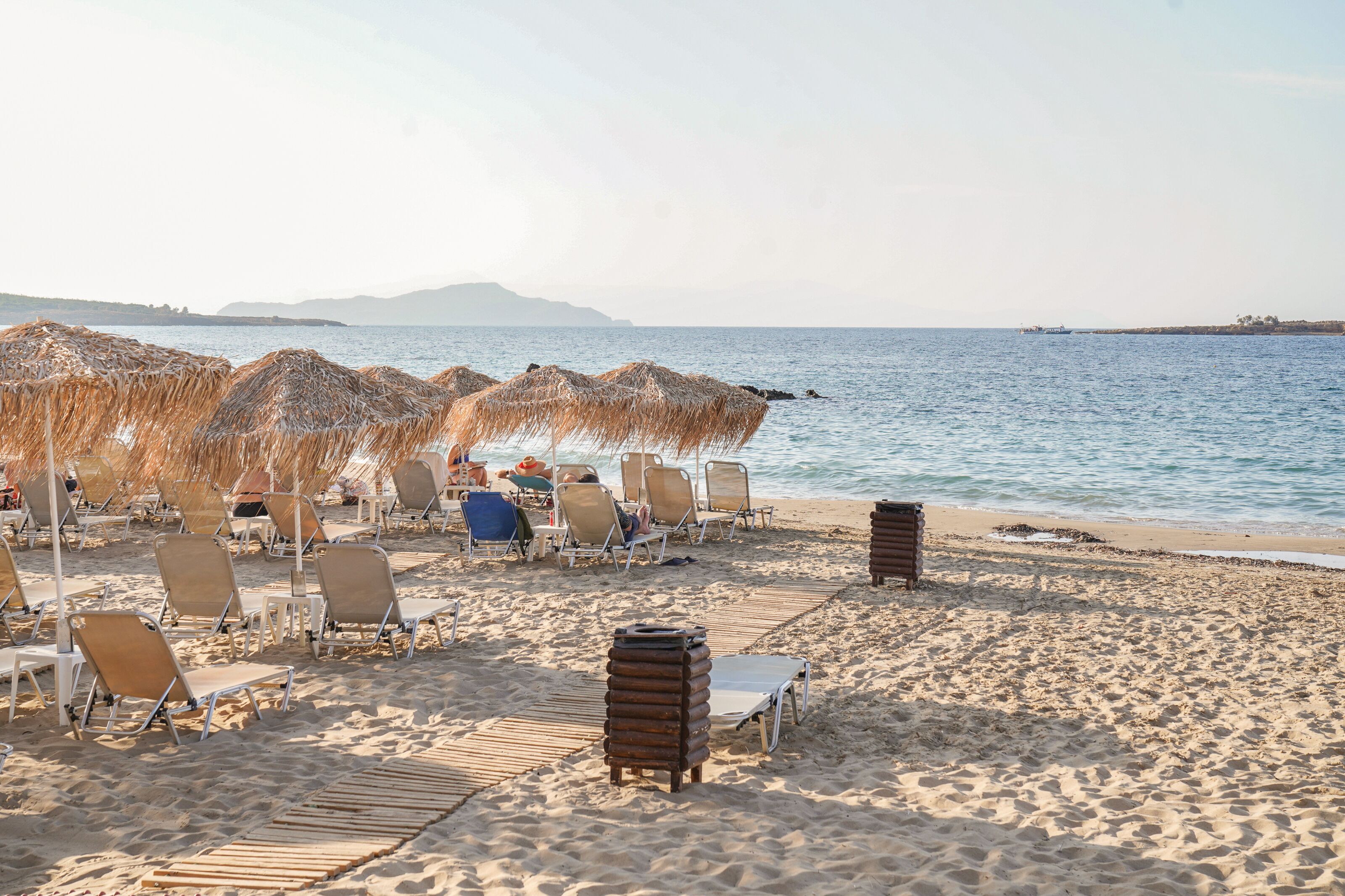 A beach with sun loungers and thatched umbrellas, a clear view of the sea, and distant mountains.
