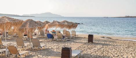 A beach with sun loungers and thatched umbrellas, a clear view of the sea, and distant mountains.