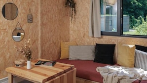 Ein gemütliches Zimmer mit Holzwand, einem fenster mit Vorhängen, einem Bett mit gestreiften und einfarbigen Kissen, einem kleinen Tisch mit einer Vase und einem Buch sowie einer hängenden Pflanze.