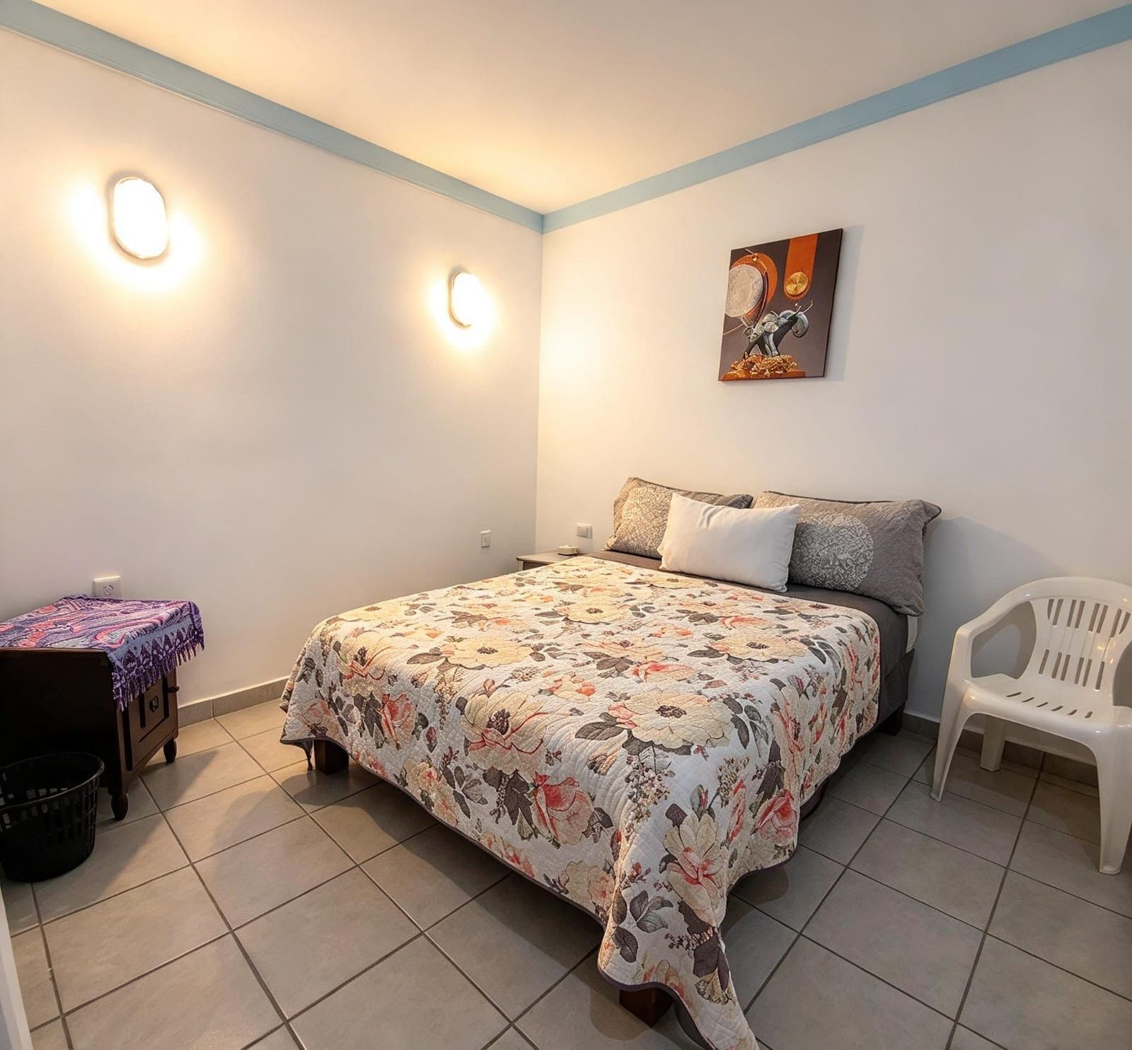 A bedroom with a bed, a chair, a small table, and a wall-mounted light fixture.