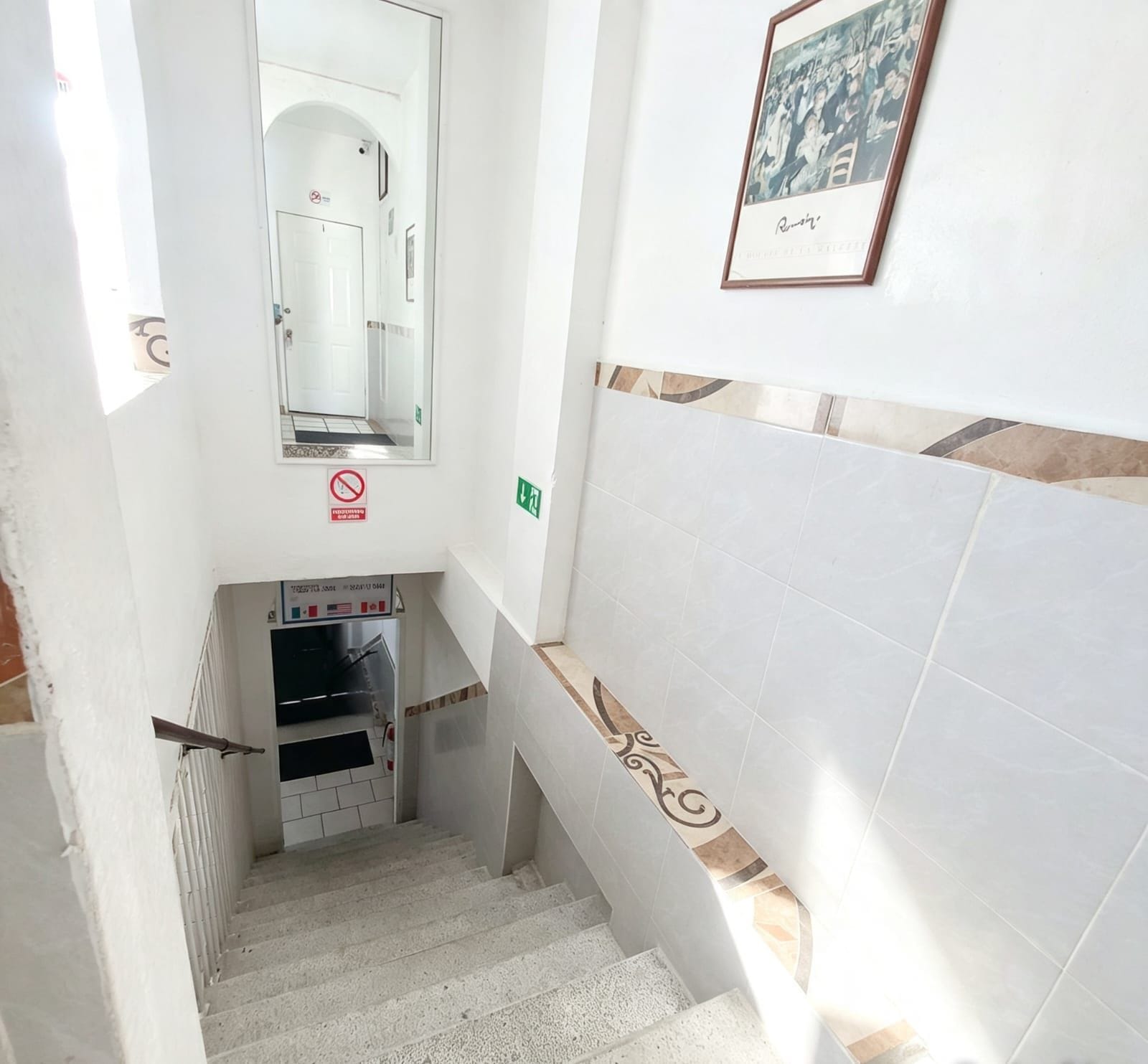 Una escalera con barandilla, un espejo arriba, una foto enmarcada en la pared y un cartel de prohibido fumar.