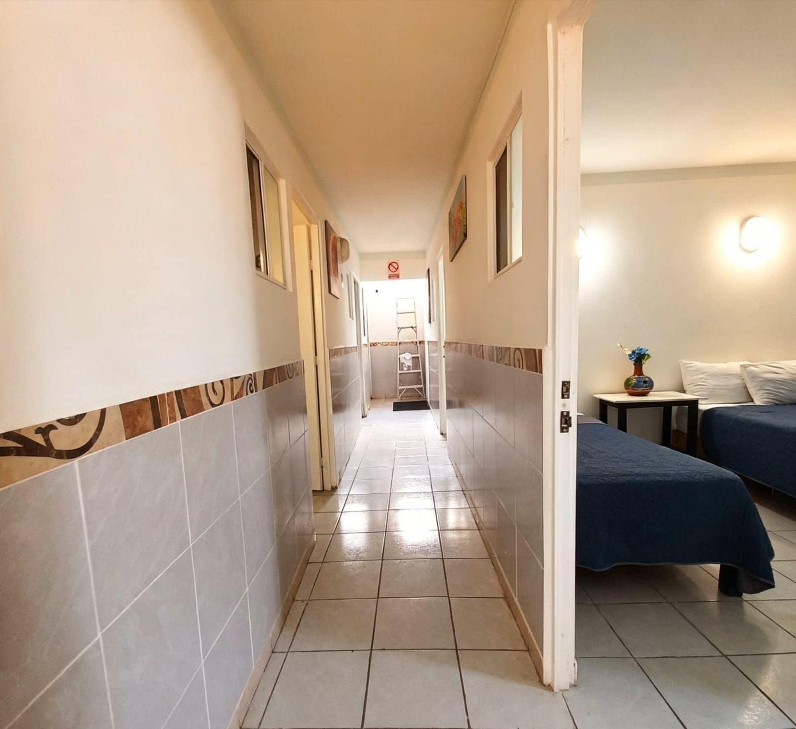 En hotellkorridor med ett klinkergolv, en säng med ett blått överkast och ett nattduksbord med en vas.