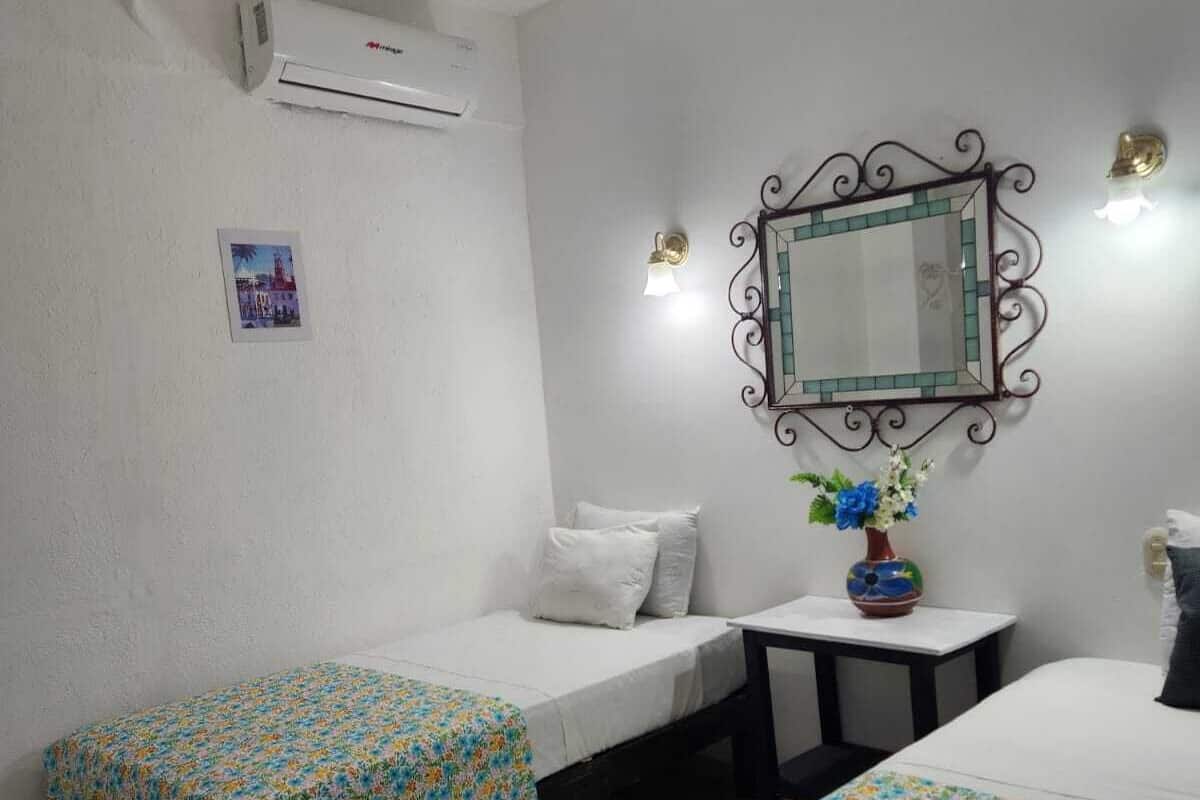 Une chambre d’hôtel avec deux lits simples, un ventilateur de plafond, un appareil de climatisation, une petite table avec un vase de fleurs et un miroir décoratif au mur.