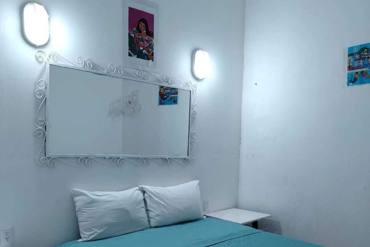 Une chambre à coucher comprenant un lit, une table de chevet, une poubelle et un miroir fixé au mur, agrémenté de lumières.