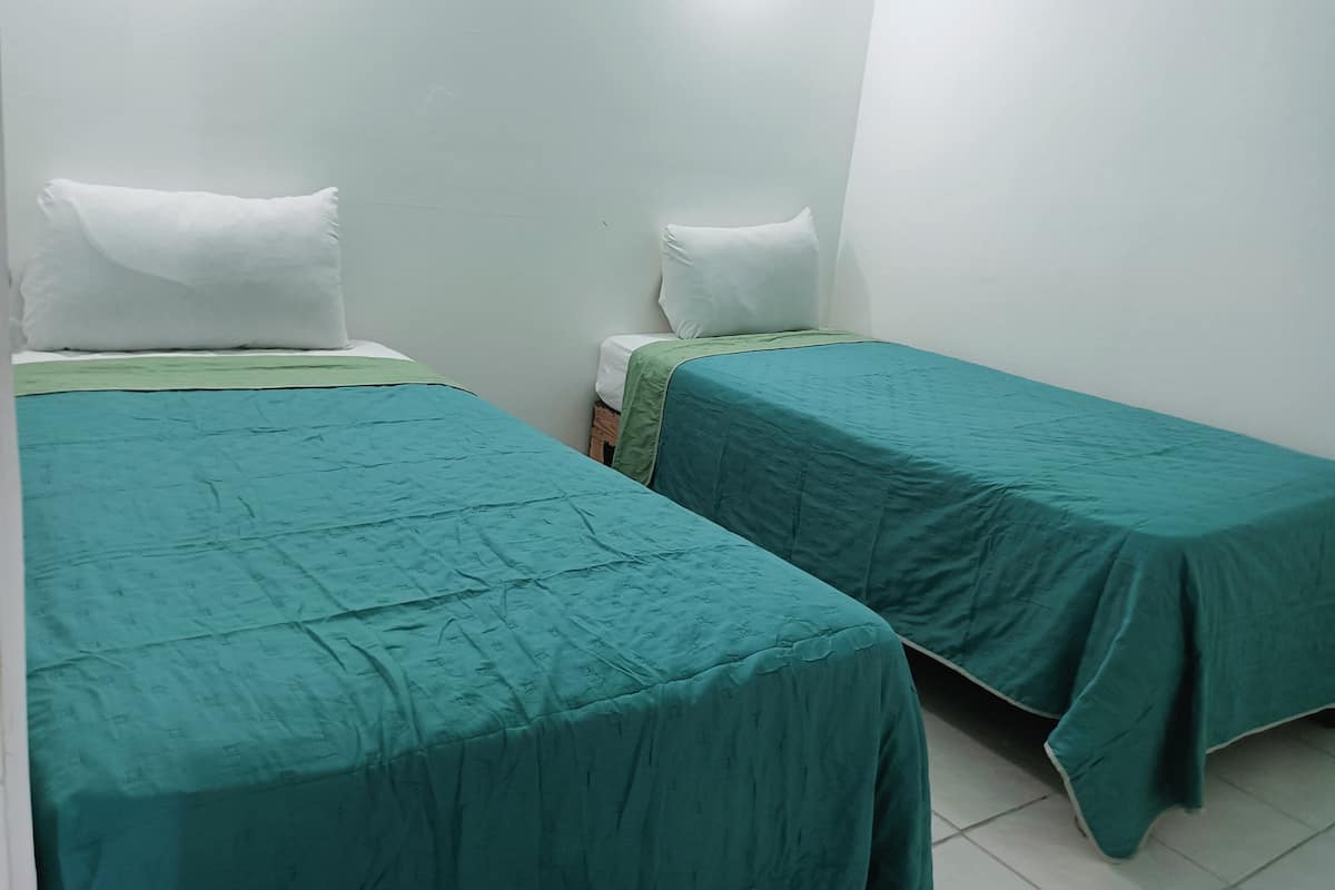 Une petite chambre propre, avec deux lits simples, chacun recouvert d’une couverture verte et agrémenté d’oreillers blancs.