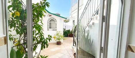 Vista a través de un ventanal hacia un patio con plantas en macetas, suelo de baldosas y un edificio con tejado y ventanas a ladrillo.