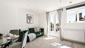 A modern living room with a green sofa, a dining table with chairs, a large window with curtains, and a radiator. - Modern Flat w/ Large Terrace | Elizabeth Line & Fast WiFi (Greater London)