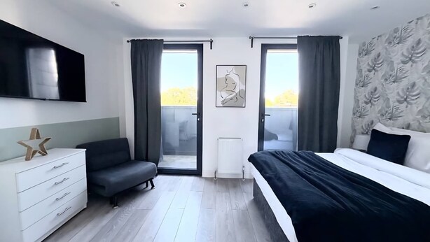 A modern bedroom with a bed, a sofa, a TV, and a balcony. - Modern Flat w/ Large Terrace | Elizabeth Line & Fast WiFi (Greater London)