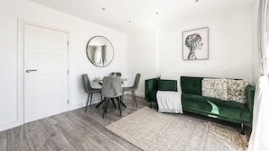 A modern living room with a dining area, a green sofa, a round mirror, and a framed abstract painting. - Modern Flat w/ Large Terrace | Elizabeth Line & Fast WiFi (Greater London)