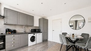 A modern kitchen with grey cabinets, a built-in washing machine, and a dining area with a round mirror. - Modern Flat w/ Large Terrace | Elizabeth Line & Fast WiFi (Greater London)