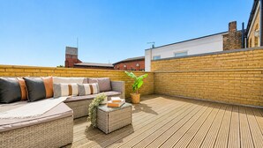 A rooftop patio with a wicker sofa, a glass coffee table, and a potted plant. - Modern Flat w/ Large Terrace | Elizabeth Line & Fast WiFi (Greater London)