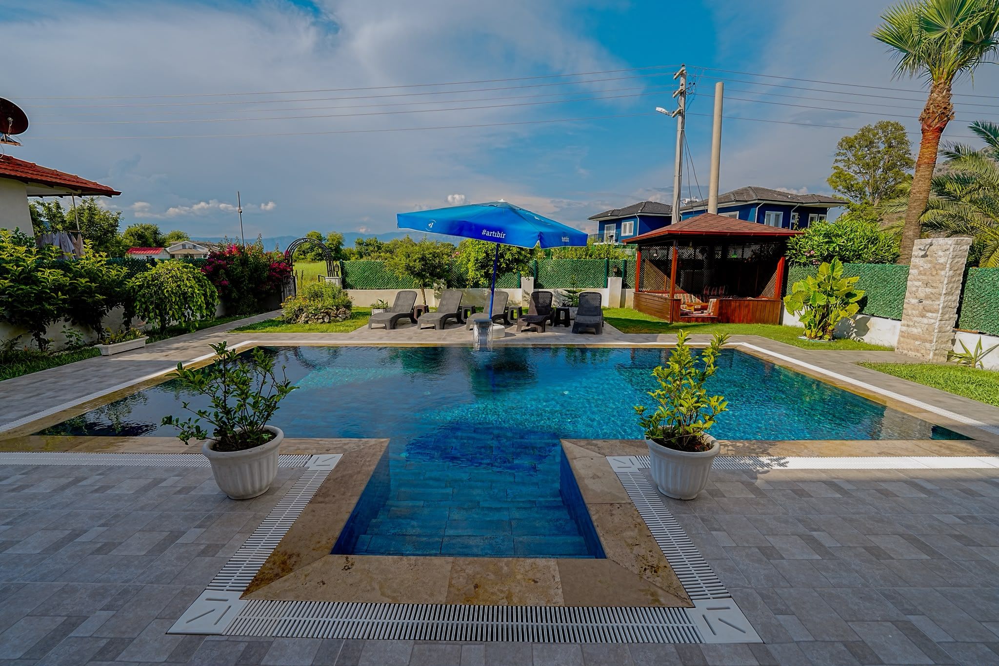 A swimming pool with lounge chairs and a gazebo, surrounded by greenery and a clear sky.