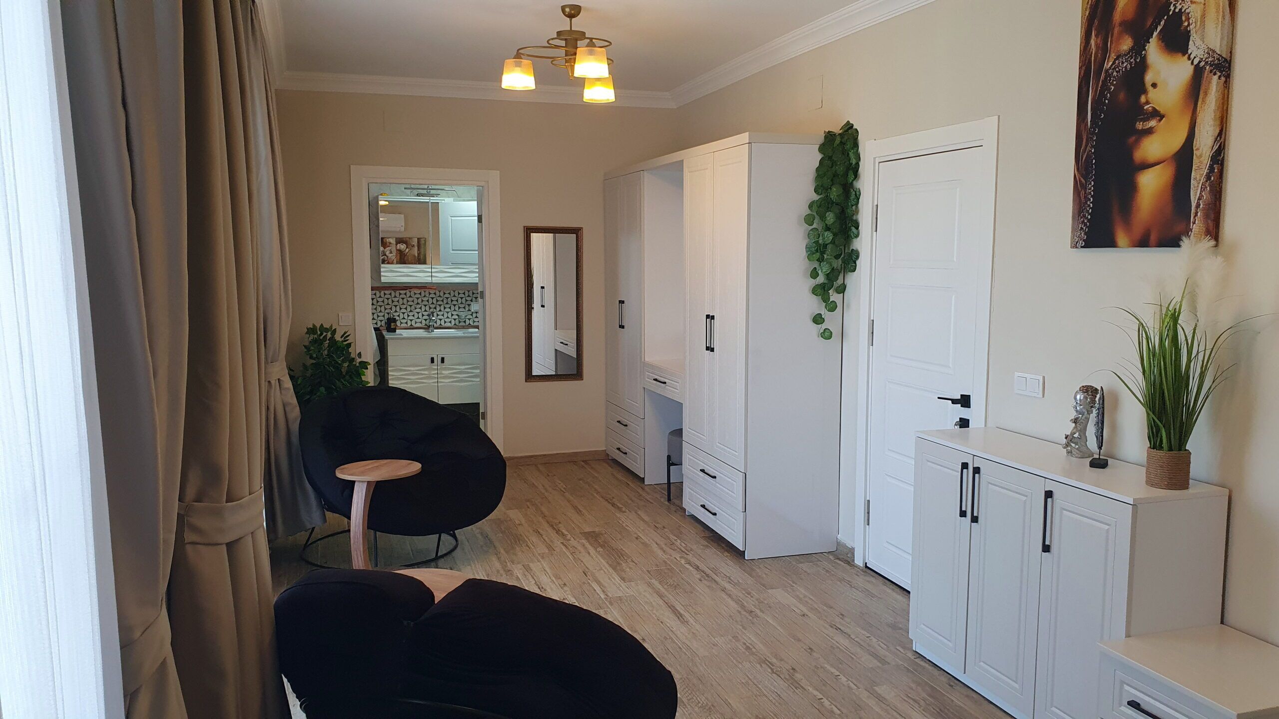 A modern apartment with a kitchen, a dining area, and a living room. There is a black chair and a small round table in the dining area. The kitchen has white cabinets and a tiled backsplash.