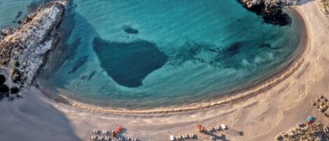An aerial view of a beach with clear blue waters, a sandy shore, and a few beach chairs and umbrellas.