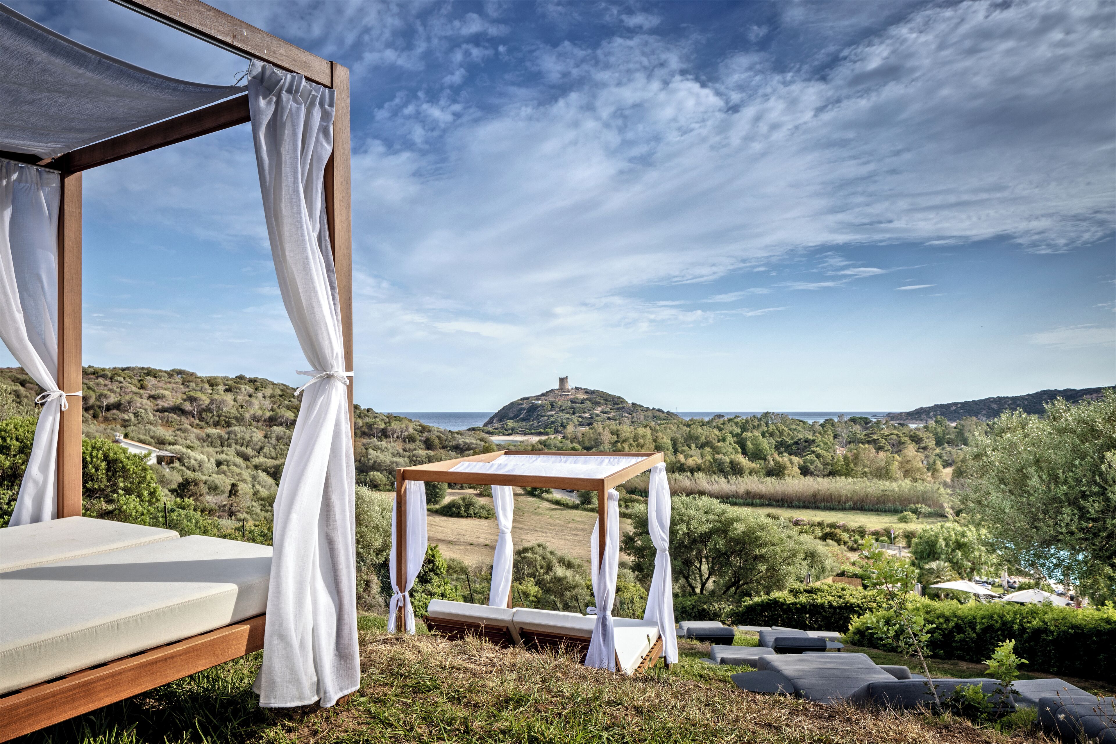 A wooden canopy bed with white curtains, a small wooden table, and a view of a hilly landscape with a distant hill and a body of water.