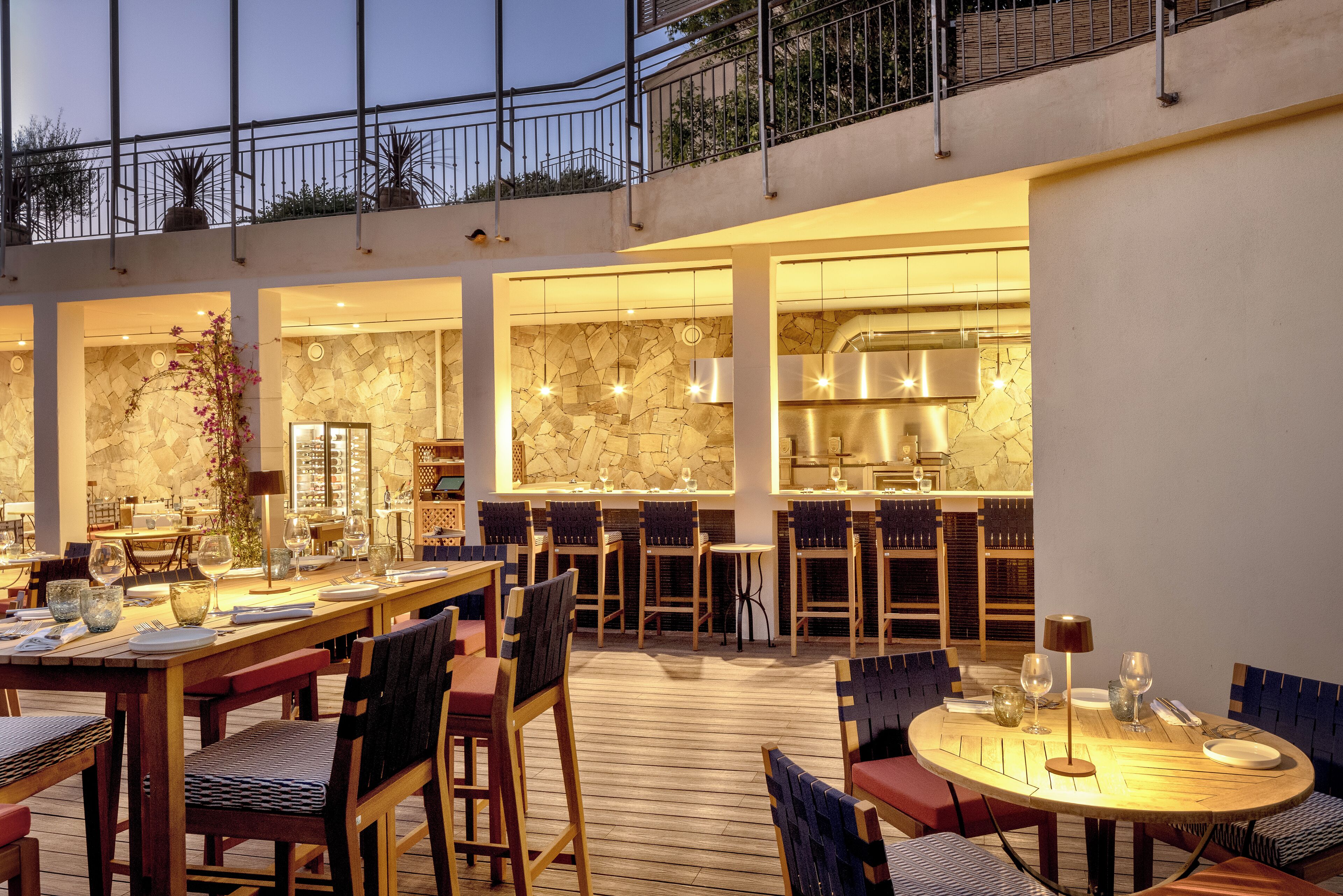 En restaurant med træterrasse, stenvægge og et køkken, der kan ses gennem store vinduer.