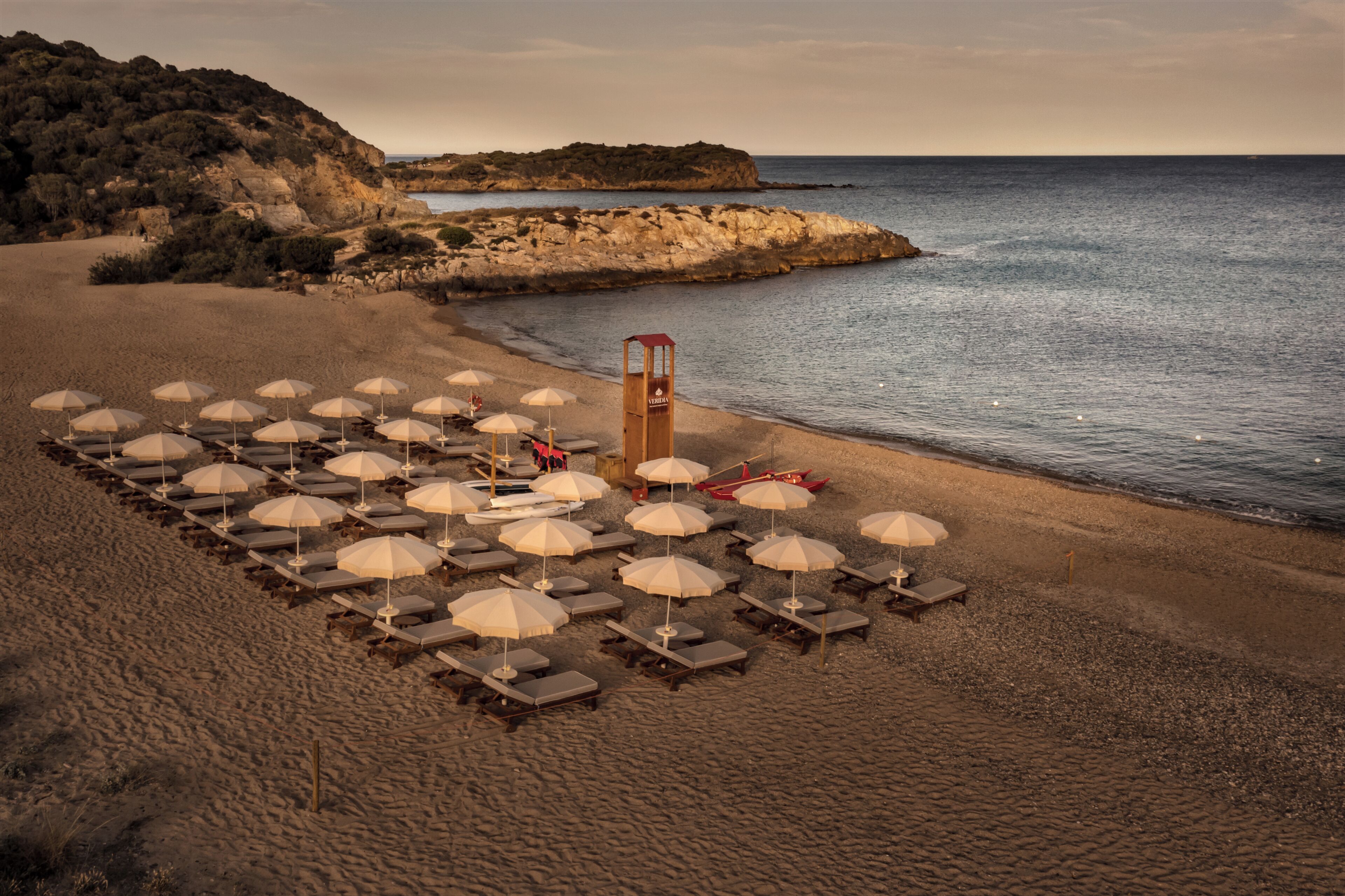 A beach with rows of sun loungers and umbrellas, a lifeguard tower, and rocky terrain in the background.