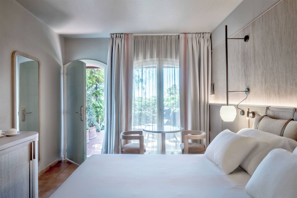 A hotel room with a large bed, a round table, two chairs, a mirror, and a view of the outdoors.