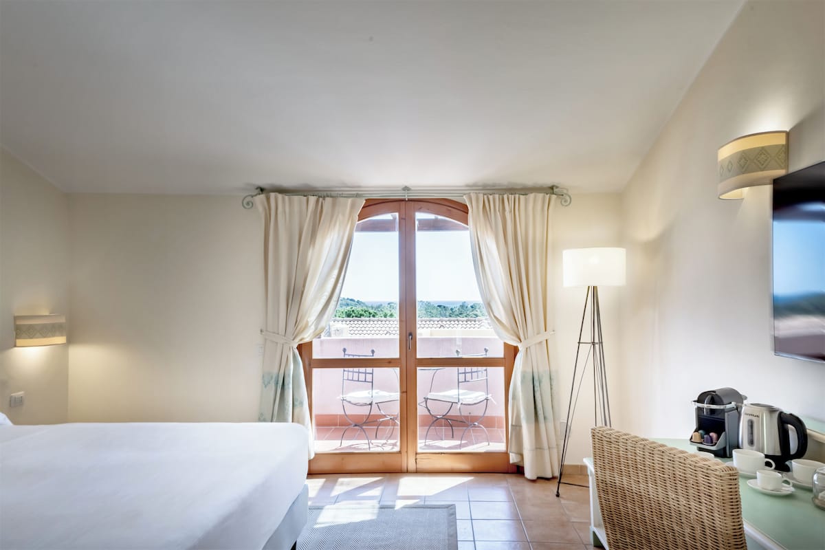 A hotel room with a bed, a dining table with chairs, a coffee maker, and a view through a glass door.