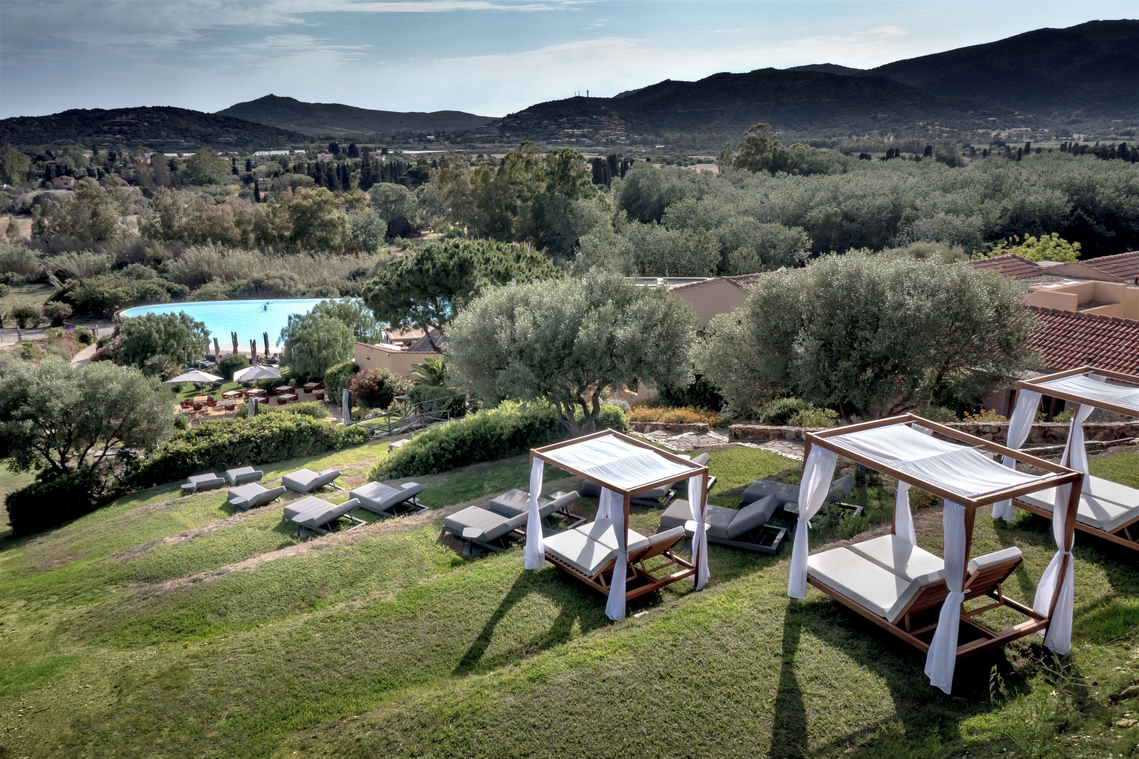Zona junto a la piscina con sillones y una mesa, rodeada de vegetación y colinas al fondo.