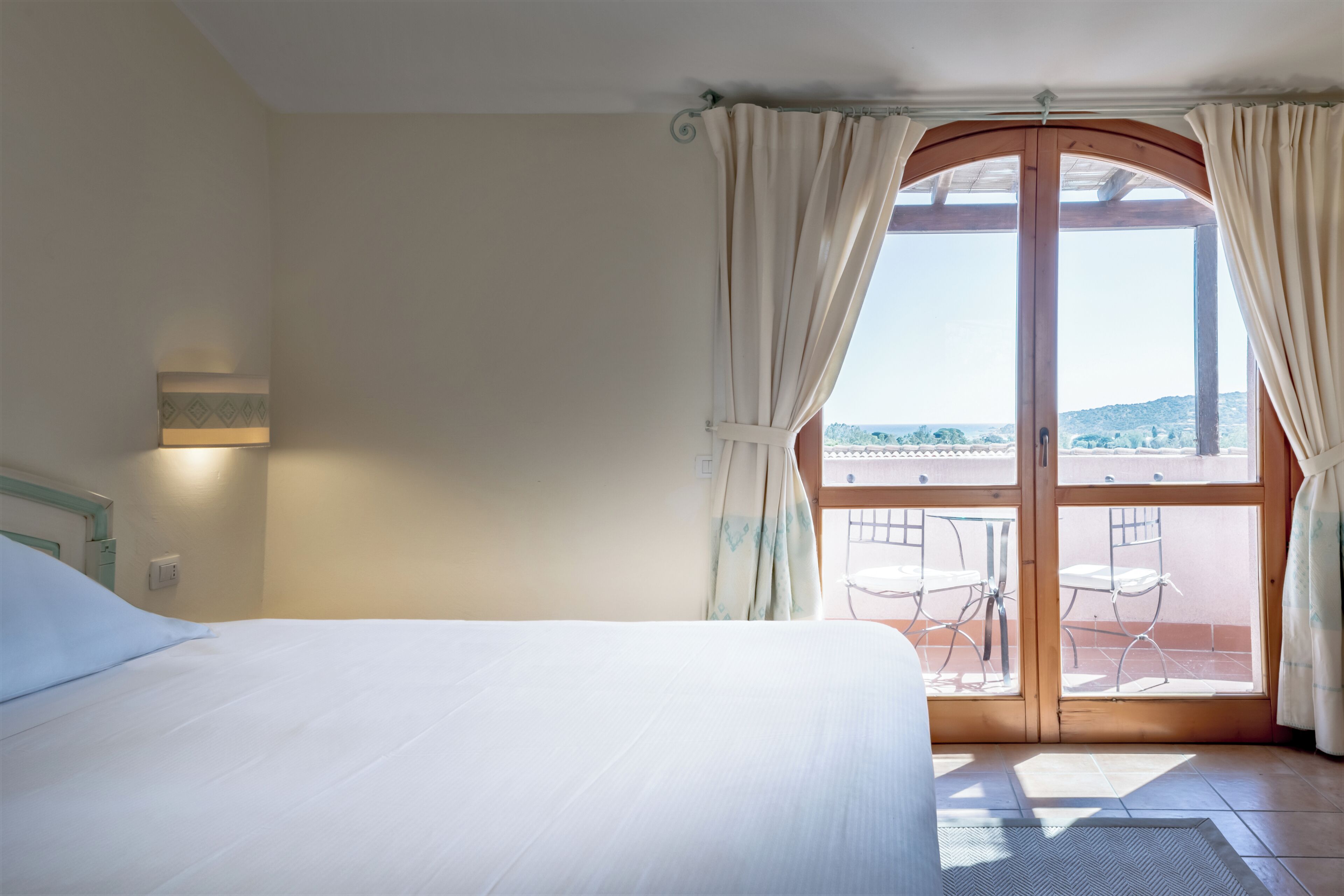 Una habitació d'hotel amb llit, finestra amb cortines, vista exterior i una taula petita amb cadires.