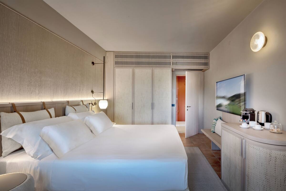 A modern hotel room with a large bed, a bedside table with a coffee maker, a TV, and a view of the city.