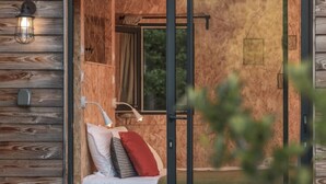 A wooden cabin with a sliding glass door leading to a bathroom with a shower, a sink, and a mirror. There is a bed with pillows and a nightstand with a lamp. - Lodge Impluvium (Volvic)